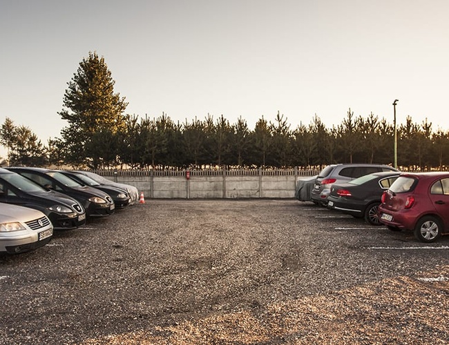 PERI Parking - Gobierno de coches aparcados en una superficie de grava. El aparcamiento está cercado y rodeado de árboles.