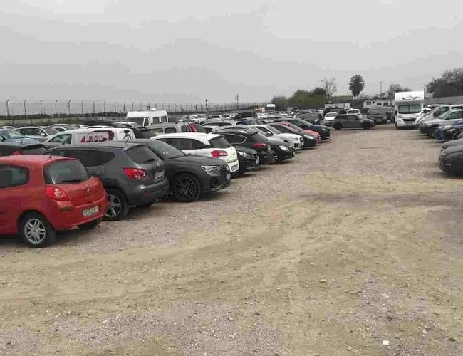 Parkovisko Drimo Park Valet - Automobily stoja v radoch na štrkovom povrchu. Areál je ohradený sieťou a obklopený zeleňou. V pozadí sú vidieť stromy a zastávka.