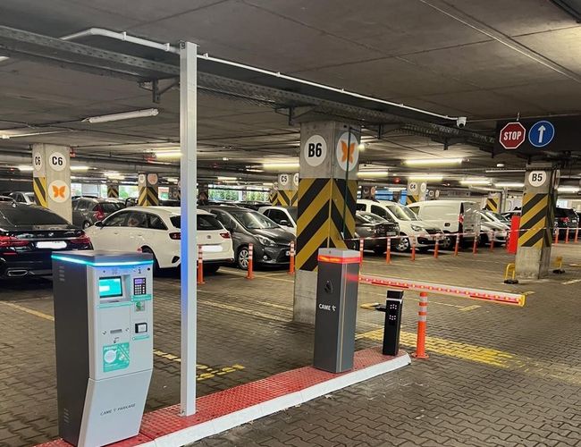 Parken P2 - Okęcie Nord Mehrstöckig - geparkte Autos und Schranke sind sichtbar. In der Mitte befindet sich ein Bezahlautomat. Wände und Säulen sind gekennzeichnet.