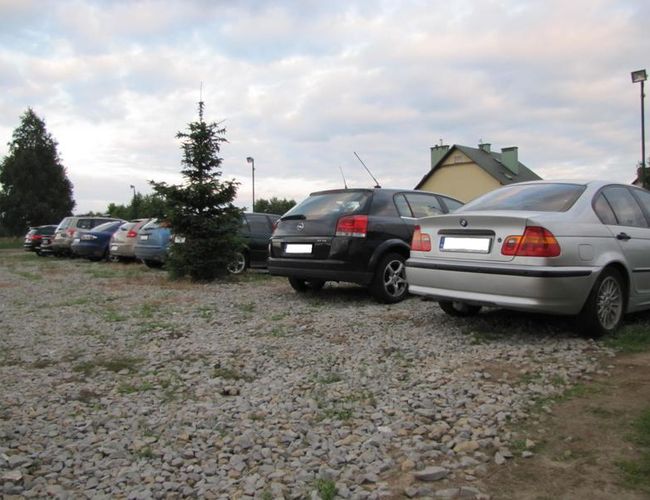 Parking Auto Park – samochody stoją na żwirowej powierzchni. Obok są drzewa i oświetlenie. W tle widać budynek.