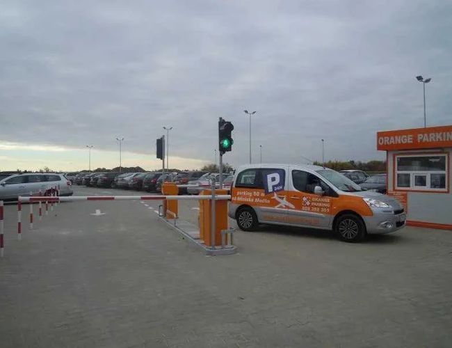 Orange Parking Modlin - El área está cercada y iluminada. Tiene una barrera de entrada y un puesto de atención. Un coche con el logo del aparcamiento está estacionado.