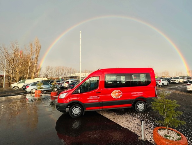 Czerwony bus do transferu pasażerów. W tle zaparkowane samochody na parkingu oraz tęcza na niebie.