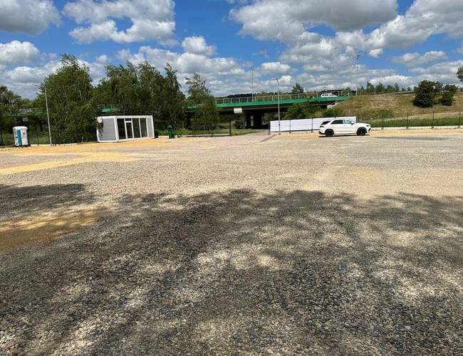 Victory Cars P2 – Estacionamiento de grava con un coche blanco. Al fondo hay un viaducto verde y árboles. Al lado hay un contenedor blanco.