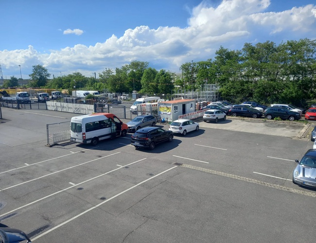 Częściowo pusty parking z kilkoma zaparkowanymi samochodami i dostawczymi autami, w tle ogrodzenie i drzewa, pod błękitnym niebem.