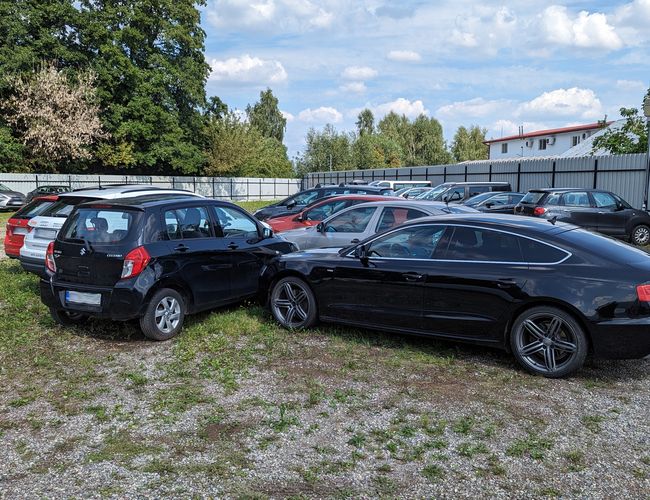 Parking P60 - Na żwirowym placu stoją zaparkowane samochody. Teren jest otoczony metalowym ogrodzeniem. W tle rośnie zieleń.