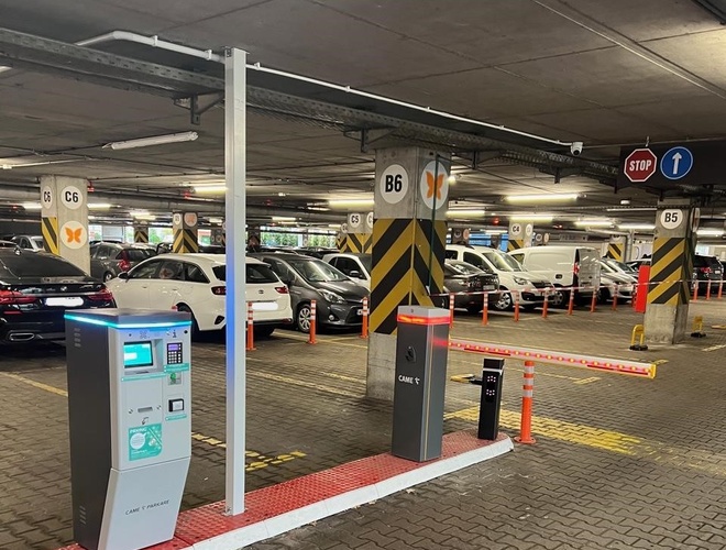 Aparcamiento P2 - Okęcie Norte Multinivel - En la foto se puede ver una fila de coches aparcados. Al fondo hay barreras de entrada y señalizaciones de lugares. El pasillo está bien iluminado.