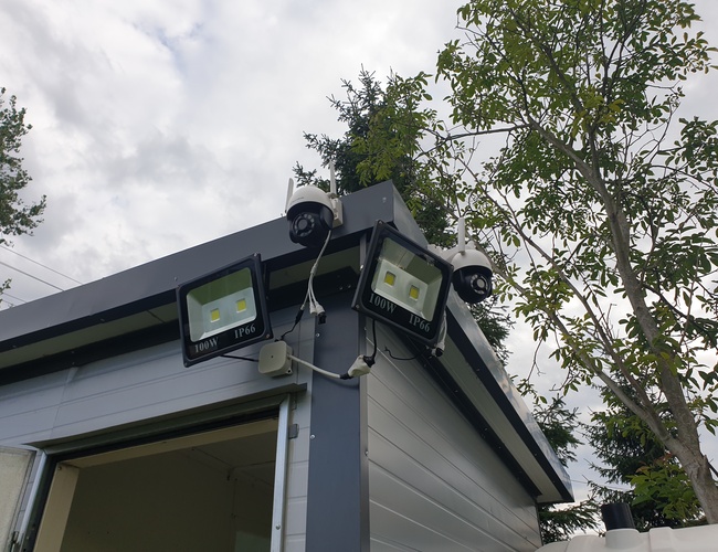 Parkovanie Park&Fly - Na obrázku je vidieť budovu s kamerami na monitorovanie. Sú tam tiež nainštalované jasné reflektory. Vedľa rastie strom.