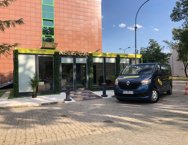 Parking Smile - Na zdjęciu znajduje się budynek recepcji. Przed nim stoi niebieski van. W tle widoczne są drzewa.