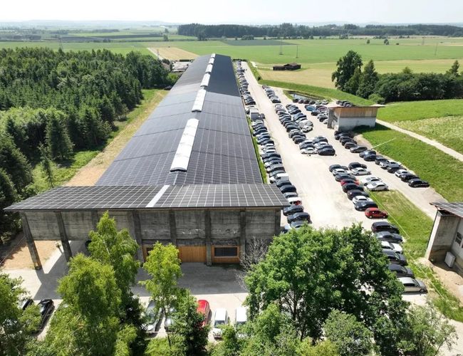 Widok z lotu ptaka na długi budynek z parkingami po obu stronach. Wokół obiektu rozciągają się tereny zielone i pola uprawne.
