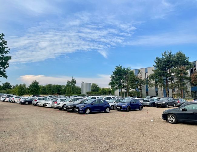 Sky Parking - Na żwirowym placu stoi wiele aut. Parking jest otoczony drzewami, a w tle widoczny jest budynek.