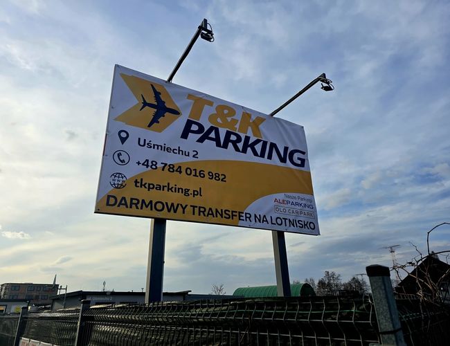 T&K Parking – Duża tablica informacyjna przy ogrodzeniu. Oferuje darmowy transfer na lotnisko. Niebieskie niebo w tle.