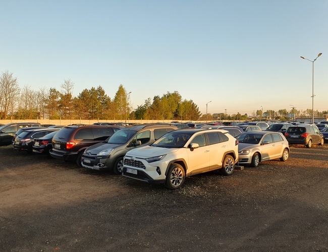 KTW Parking - Na żwirowym placu stoją zaparkowane samochody. Teren jest otoczony ogrodzeniem i oświetlony latarniami.