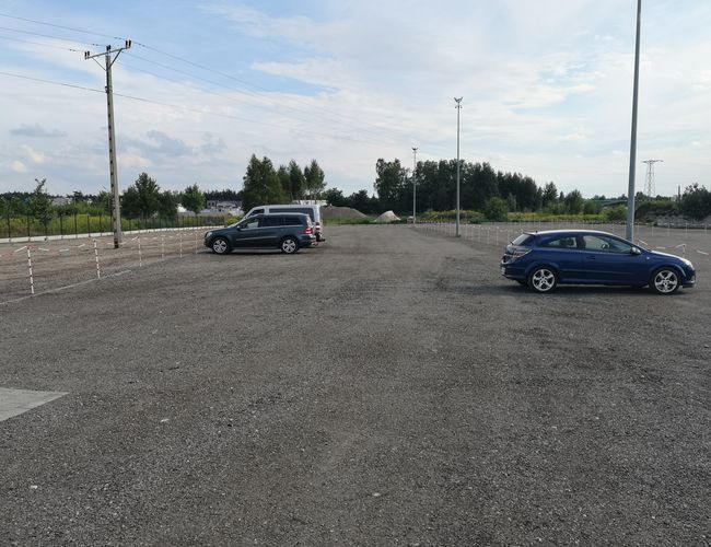 Parking A1 Rondo - Na żwirowym placu stoją dwa samochody. Teren jest ogrodzony i oświetlony. Widzimy drzewa i linie elektryczne.