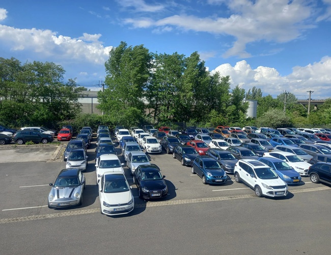 Parking z wieloma rzędami samochodów osobowych, częściowo zacieniony przez pobliskie drzewa, widok z góry.