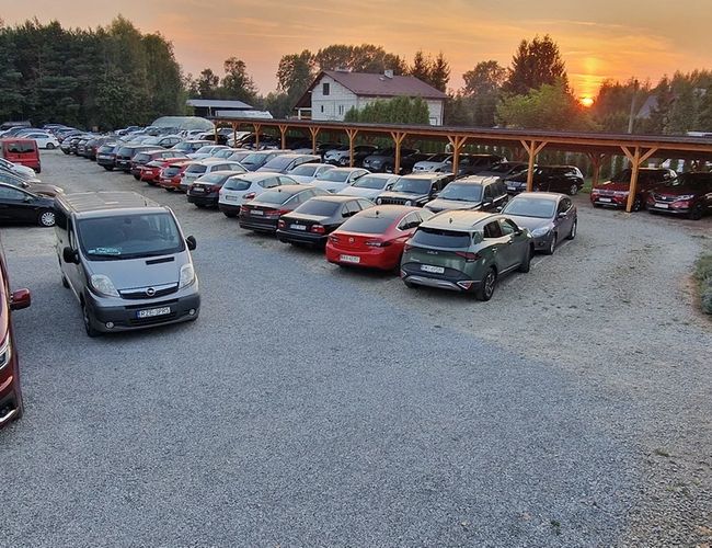 Aparcamiento PARK4YOU - En el patio de grava hay muchos coches. Parte de los coches está bajo el techo. Al fondo se puede ver el atardecer y la vegetación.