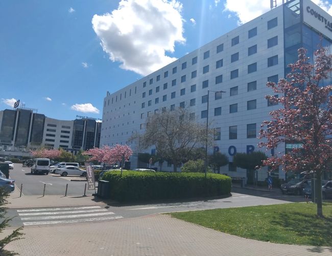 PRG Parking – Na zdjęciu widać przestronny parking. Obok stoją wysokie biurowce. Drzewa wokół są kwitnące.