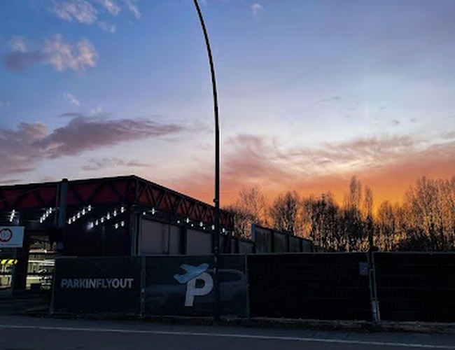 Parkovisko Park In Fly Out – oplotený pozemok s lampou. Na pozadí je vidieť zapadajúce slnko a stromy. Nebo je v farbách západu.