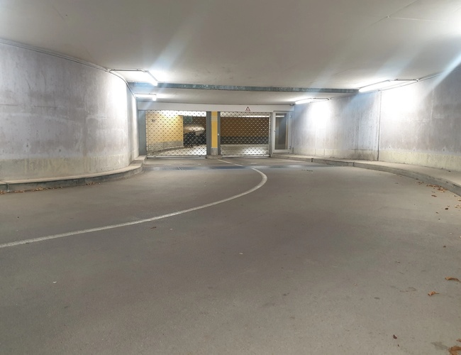 Zakręt wjazdowy na podziemny parking, betonowe ściany i jasne oświetlenie.