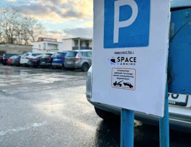 Parkovanie na parkovisku – Na parkovisku stojí značka označujúca miesto na parkovanie. Na pozadí sú viditeľné zaparkované autá a budova. Je slnečno.