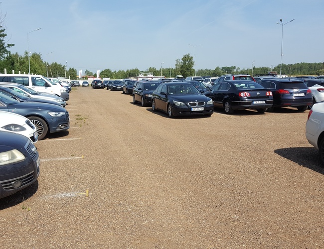 AAP-66 – Parking z dużą ilością aut. Teren jest nieutwardzony, z oznaczeniami miejsc. Po lewej stronie jest ogrodzenie.