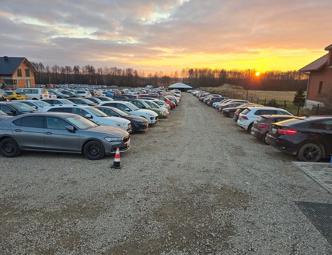 AleParking – szutrowy parking pełen samochodów. Otoczony budynkami i drzewami. Słońce zachodzi na horyzoncie.