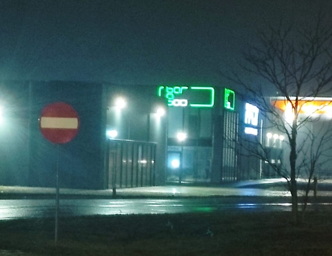 Q-Parking — Na pozadí neónový nápis nad budovou. V popredí cesta s zákazom vjazdu. Večerná scénka s mestským osvetlením.