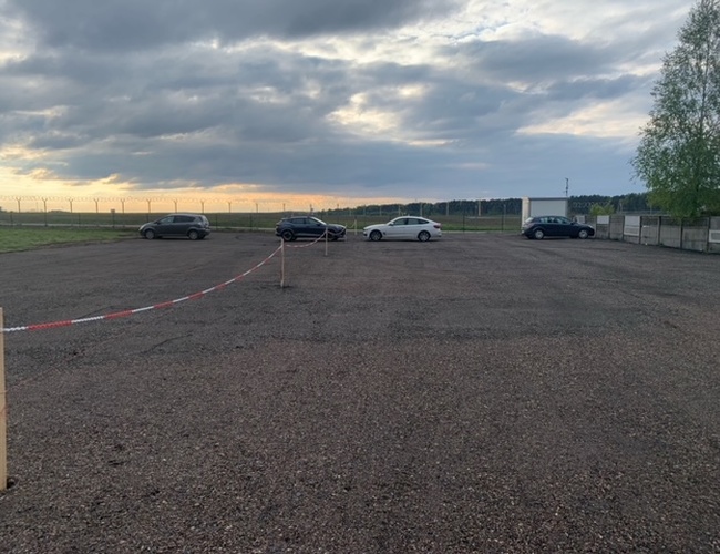Aparcamiento con vista – un área de estacionamiento con algunos coches. El terreno está abierto y sin iluminación. En el fondo se puede ver una cerca y árboles.