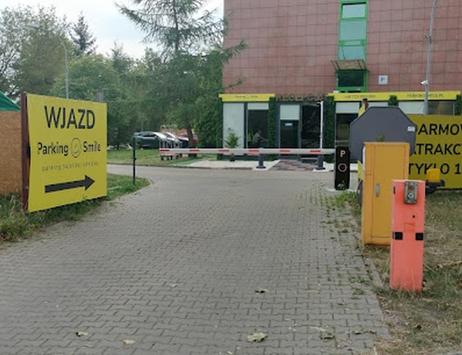 Parking Smile - Wjazd oznaczony żółtymi tablicami. W tle budynek z bramą. Teren jest ogrodzony.