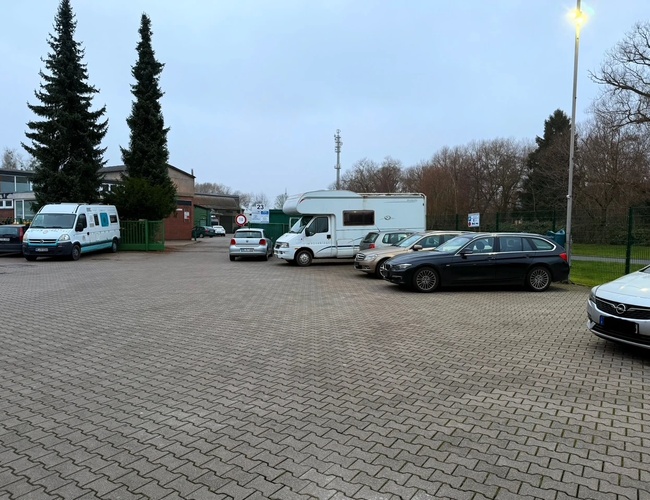 Parking Park Elite Valet – Na parkingu stoi kilka samochodów. Teren jest wyłożony kostką i ogrodzony. W tle widać drzewa i budynek.