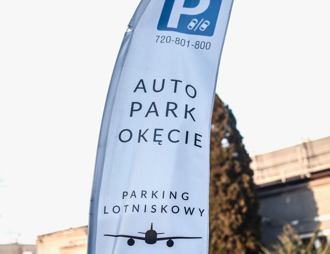 Auto Park Okęcie – Reklamná vlajka pri vstupe na parkovisko. V pozadí je vidieť čierne auto a budova.
