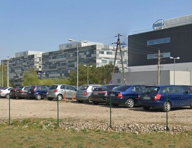 Parkovisko Piątka - Parkovisko s oplotením a stojacimi autami. Na pozadí moderné budovy.