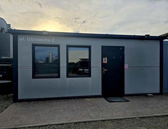 T&K Parking – Šedá kancelária s dvomi oknami a dverami. Dvere majú značku zákazu fajčenia. Pred kanceláriou sú dlažobné kocky.