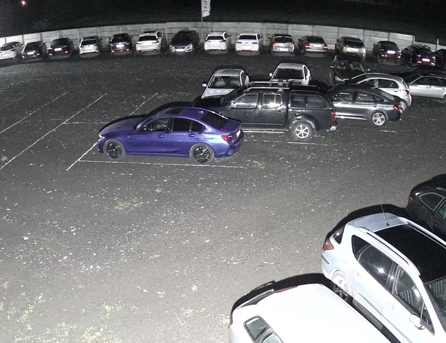 TRIP Parking - Nočné, dobre osvetlené parkovisko. Automobily stojí zaparkované blízko seba na asfaltovom povrchu.