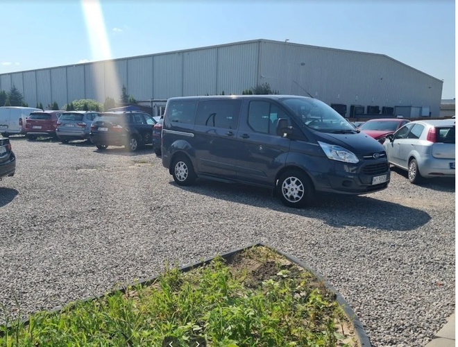 Aparcamiento Dominik - El terreno es de grava y no está cercado. Varios vehículos están estacionados. Al fondo se puede ver un gran edificio.