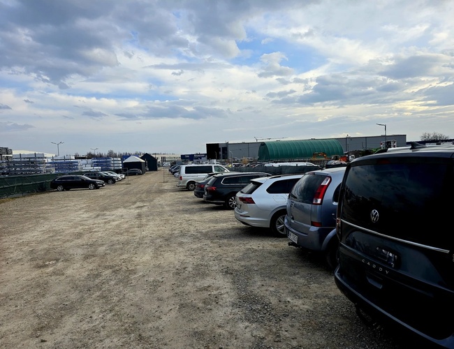 T&K Parkovisko - Riad zaparkovaných áut na štrkovej ploche. Pozemok je obklopený ohradou. Na pozadí je vidieť veľkú budovu.