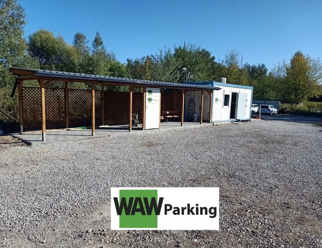 Parkovanie WAW – Štrkovisko s pristrešením a kontajnerom. Vedľa rastú stromy a stoja autá. Počasie je slnečné.