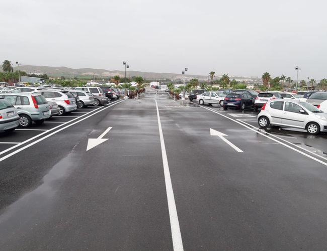 Victoria Parking Walencja - parking z wieloma samochodami. Równe linie oznaczają miejsca parkingowe. W oddali widoczne są palmy i wzgórza.