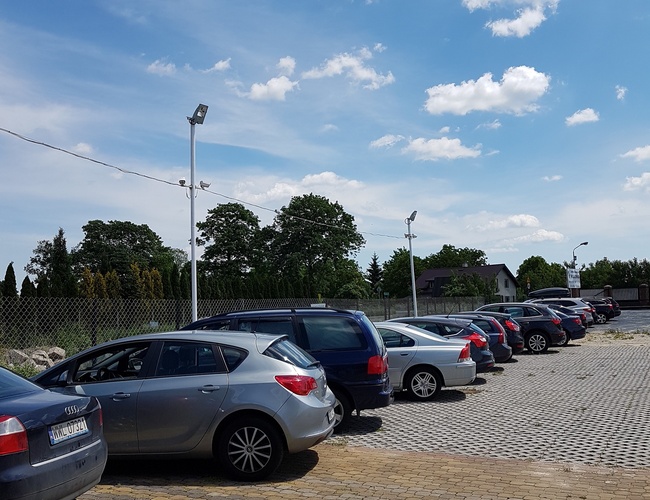 PARKING P24 PREMIUM - Samochody stoją na utwardzonym placu. Teren jest ogrodzony i oświetlony. Na niebie widać kilka chmur.