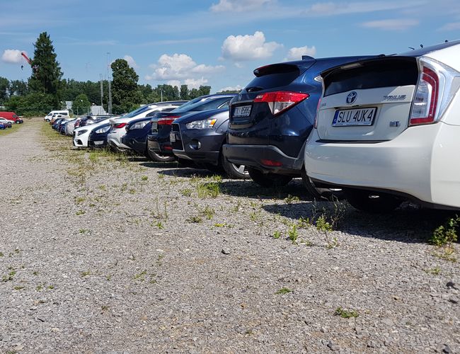 GOOD Parking - Szutrowy teren z zaparkowanymi samochodami po obu stronach. W tle widać drzewa i niebo z chmurami.