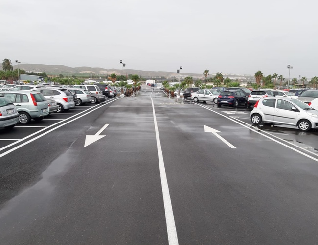 Victoria Parking Walencja - parking z wieloma samochodami. Równe linie oznaczają miejsca parkingowe. W oddali widoczne są palmy i wzgórza.