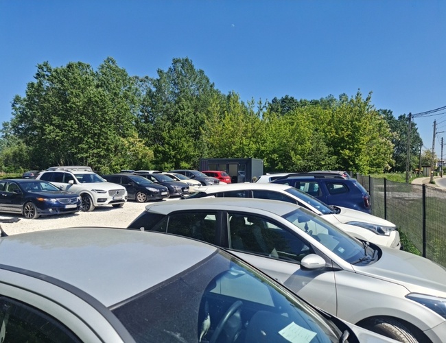 MyParking – Pozemok je ohradený a čiastočne tieni stromami. Na parkovisku stojí veľa áut rôznych značiek a farieb.
