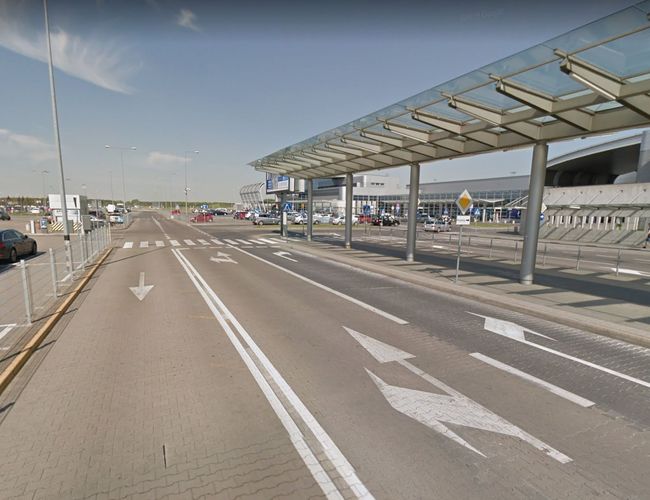 Oficjalny parking P2 - Droga prowadzi wzdłuż zadaszonego chodnika. Obok znajdują się zaparkowane samochody i budynek z napisami.