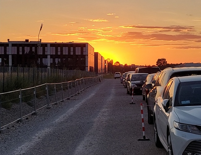 Holiday Parking - Rząd samochodów stoi wzdłuż żwirowej drogi. Obok drogi jest ogrodzenie. W tle zachodzi słońce za nowoczesnym budynkiem.