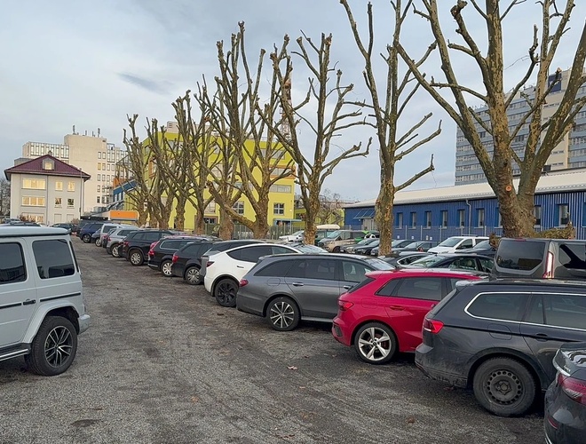 Parken Park & Go Valet – Platz mit kieseligem Untergrund. Umzäunt mit einem Zaun, sichtbare geparkte Autos. Rundherum Gebäude und Bäume.