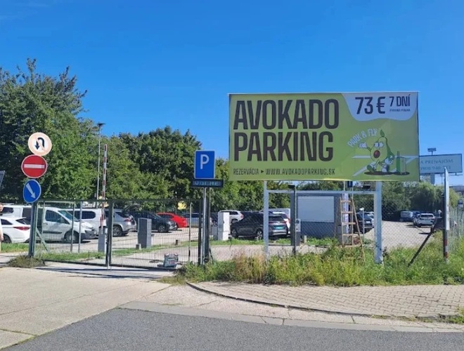 Tablica reklamowa „Avokado Parking” przy wjeździe na parking, obok drogi i zaparkowanych samochodów, w słoneczny dzień
