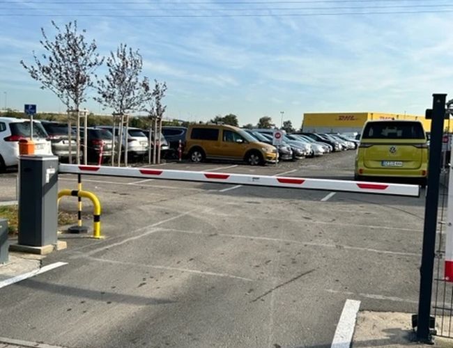 Parking P365 Valet - Na parkingu są zaparkowane samochody. Wjazd blokuje szlaban. Obok rosną małe drzewka.