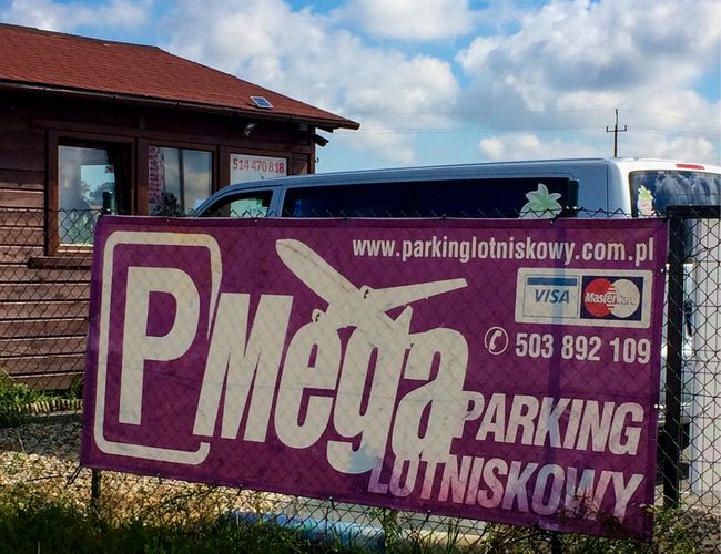 Parkplatz Mega Park Service - Neben dem Gebäude steht ein Auto. Ein Zaun mit Parkplatzwerbung. Es ist sonniges Wetter.