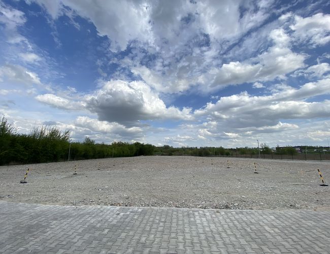 SkyWay Parking – duży utwardzony plac z kostki. Teren jest ogrodzony i otoczony zielenią. Na niebie są białe chmury.
