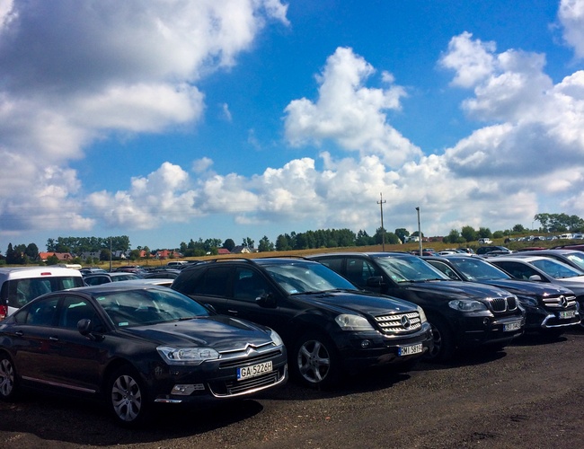 Parkovisko Mega Park Service - Na parkovisku stojí veľa osobných áut. Je slnečný deň s miernym oblačnosťou. Pozemok nie je oplotený.