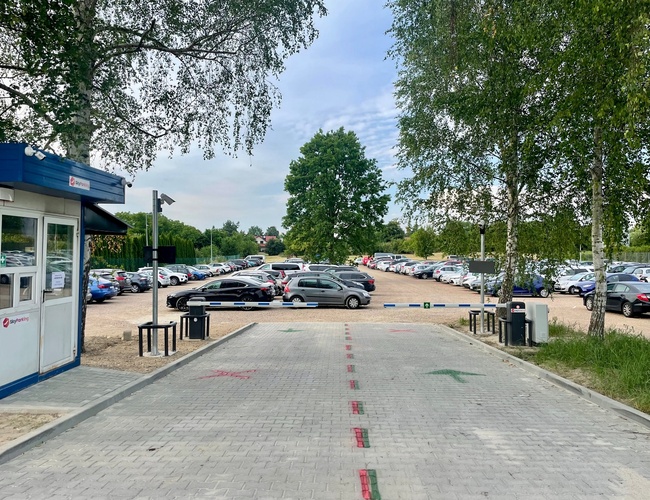 Sky Parking – Parkovisko je oplotené a plné áut. Vjazd je spevnený a osvetlený lampami. Je vidieť malú budovu pri vstupe.